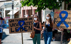 Zwei Frauen mit medizinischen Masken und Transparenten auf einer Demo für bessere Forschung und Versorgung von ME/CFS Patient:innen