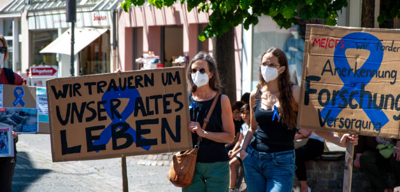 Zwei Frauen mit medizinischen Masken und Transparenten auf einer Demo für bessere Forschung und Versorgung von ME/CFS Patient:innen